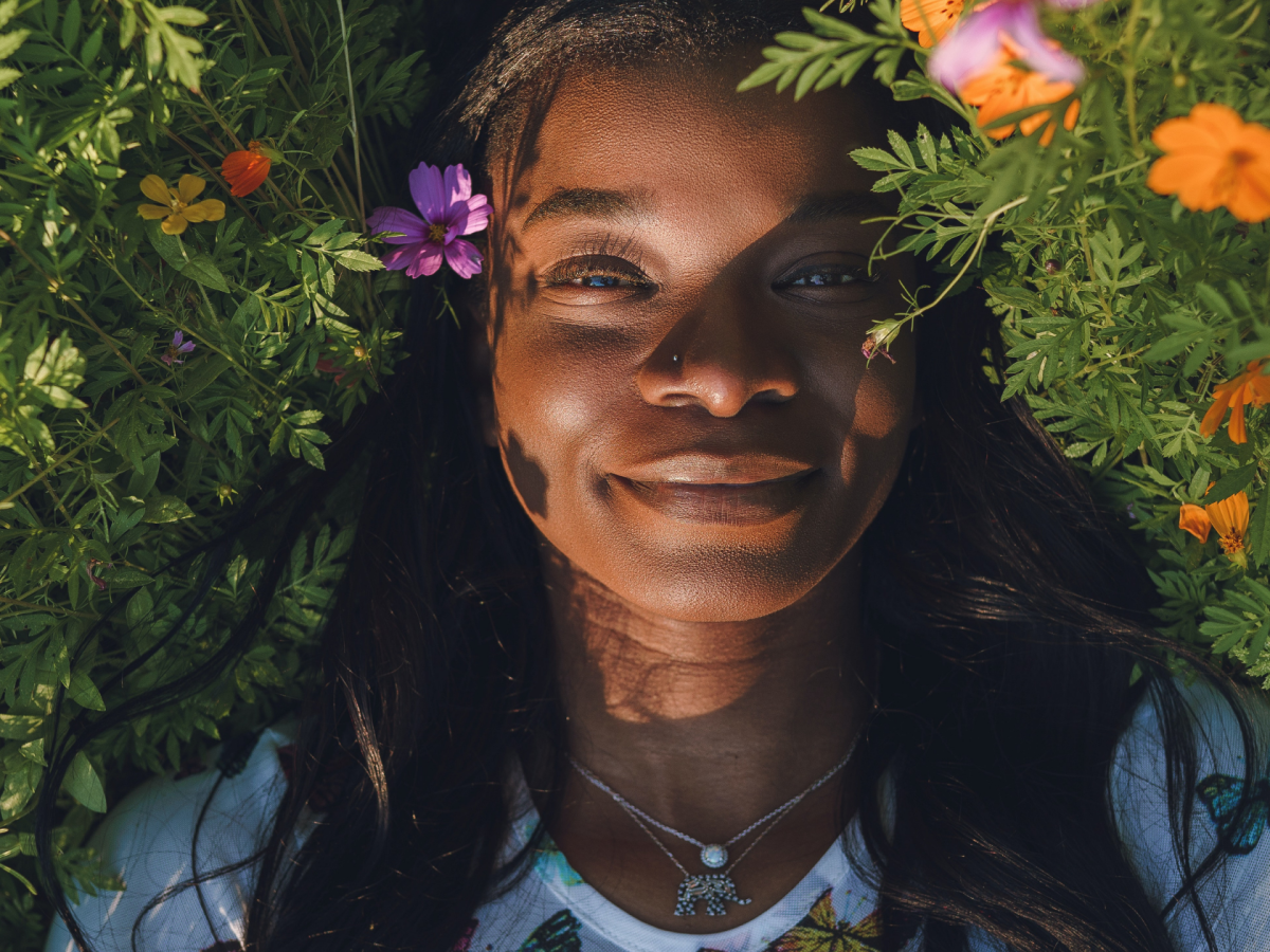 A dark skin black person smiling while laying on grass with various flowers. they have long straight hair and a nose ring and are wearing a butterly shirt